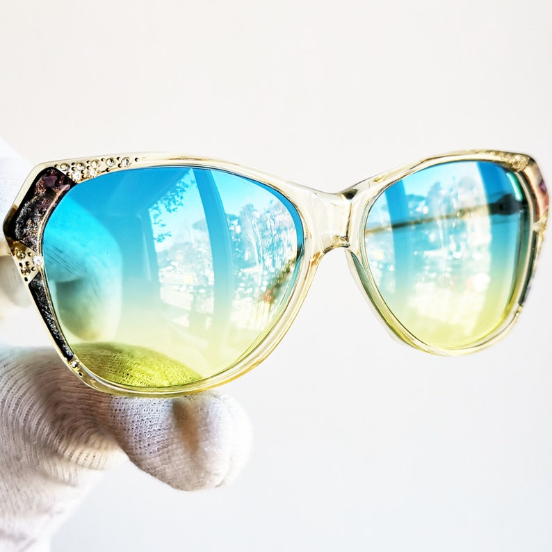 May include: Clear-framed sunglasses with blue and yellow gradient lenses. The frames have decorative detailing on the upper corners. The lenses reflect a bright outdoor scene. A stylish accessory for eye protection.