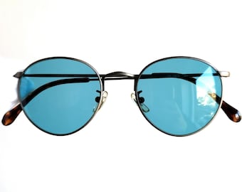 May include: Round sunglasses with a dark metal frame and blue lenses. The temples have a tortoiseshell pattern. The sunglasses are set against a white background.