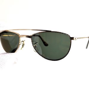 May include: Gold-tone sunglasses with green lenses. The sunglasses have a double bridge and a patterned design on the frame. The temples are thin and gold-colored.