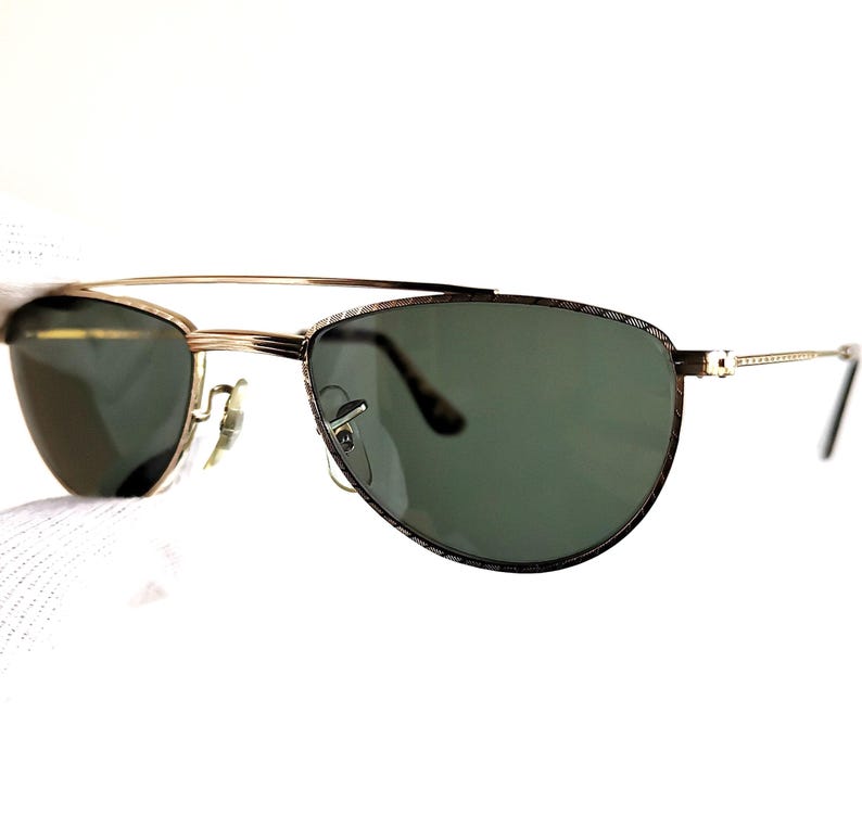 May include: Gold-tone sunglasses with green lenses. The sunglasses have a double bridge and a patterned design on the frame. The temples are thin and gold-colored.