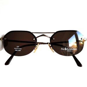 May include: Brown sunglasses with a gold frame and brown lenses. The sunglasses have a label that says "Polo Ralph Lauren Eyewear" and "UV Protected 400 NM".