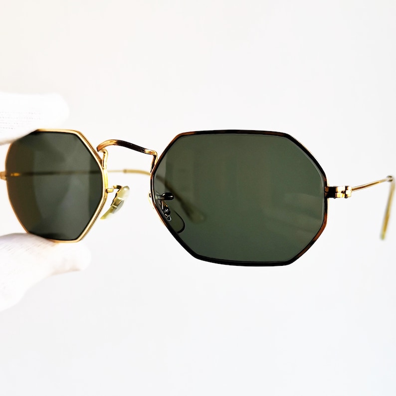 Pu&ograve; includere: Occhiali da sole con montatura esagonale dorata e lenti verde scuro.