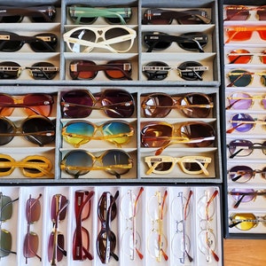 Puede incluir: Una colecci&oacute;n de gafas de sol y gafas graduadas en varios estilos y colores, expuestas en bandejas organizadas. El surtido incluye monturas con detalles en negro, marr&oacute;n y dorado, as&iacute; como lentes de colores.