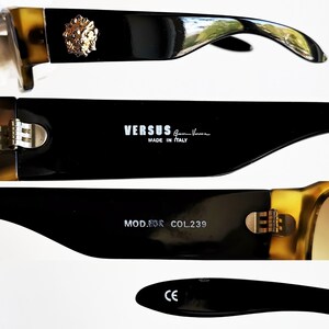 May include: Black and brown sunglasses with gold accents. The sunglasses feature a Versace logo and the text 'VERSUS Gianni Versace MADE IN ITALY' on the side. The sunglasses also have the text 'MOD. 8008 COL.239' and 'CE' on the arms.