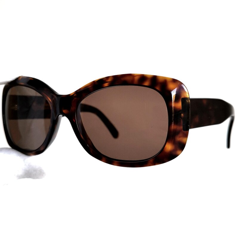 Puede incluir: Gafas de sol carey con lentes marrones. Las monturas tienen una forma rectangular redondeada. Las patillas son de color marr&oacute;n oscuro y se curvan ligeramente en los extremos. Las gafas de sol est&aacute;n sobre un fondo blanco.