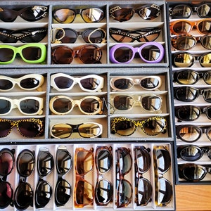 May include: A collection of 10 rows of sunglasses in various colours and styles, including black, brown, green, blue, and white. The sunglasses are arranged in a grid pattern on a white background.