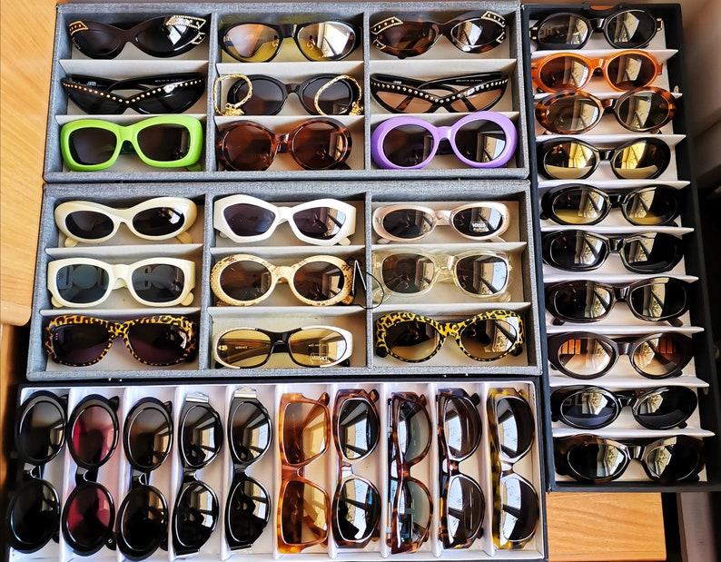 May include: A collection of 10 rows of sunglasses in various colours and styles, including black, brown, green, blue, and white. The sunglasses are arranged in a grid pattern on a white background.