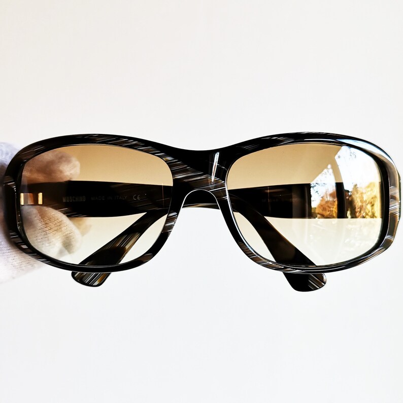 Puede incluir: Gafas de sol Moschino negras y marrones con montura rectangular y lentes degradadas. Las gafas de sol tienen un patr&oacute;n de rayas &uacute;nico en la montura y detalles dorados. Fabricadas en Italia.