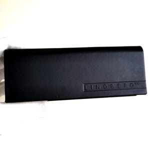 May include: A rectangular black hard-shell glasses case with a smooth, matte finish. The case features a horizontal embossed line detail across the lower portion, with the brand name LINDBERG embossed in capital letters on the bottom right side.