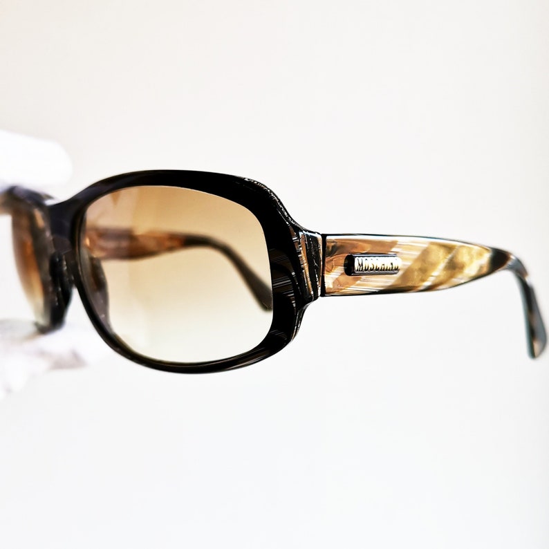 Puede incluir: Gafas de sol negras y marrones con lentes degradadas. Las monturas tienen un patr&oacute;n &uacute;nico y el nombre de la marca "MOSCHINO" es visible en la patilla. Estas gafas ofrecen protecci&oacute;n ocular y estilo.