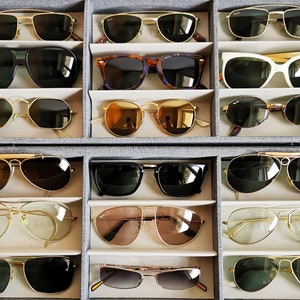 Pu&ograve; includere: Una collezione di 12 paia di occhiali da sole esposti in una vetrina bianca e grigia. Gli occhiali da sole sono disposti in file di tre, con ogni fila che presenta uno stile diverso di occhiali da sole. Gli occhiali da sole sono tutti di vari colori, tra cui nero, marrone, oro e bianco.