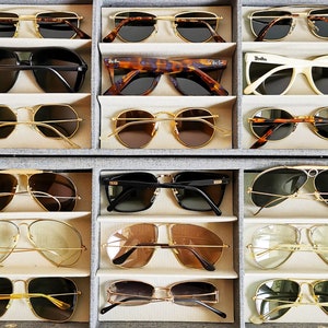 May include: A collection of sunglasses in various styles and colors, including aviator, round, and square frames. The sunglasses are displayed in a gray organizer, showcasing a variety of lens tints and frame materials, such as gold and black.