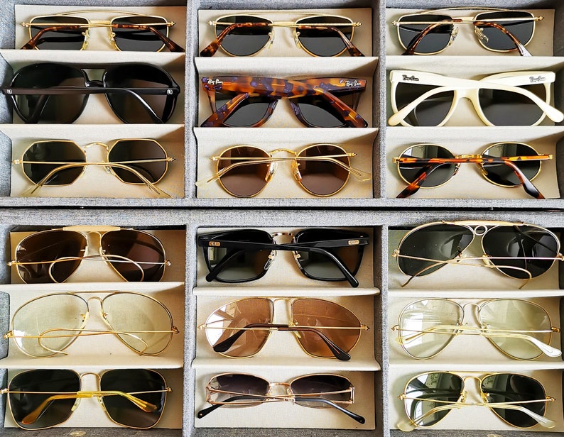 May include: A collection of sunglasses in various styles and colors, including aviator, round, and square frames. The sunglasses are displayed in a gray organizer, showcasing a variety of lens tints and frame materials, such as gold and black.