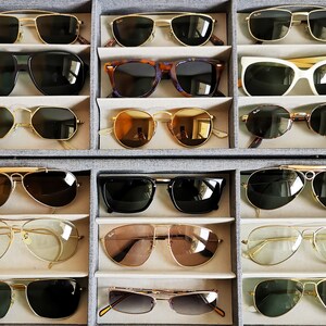 May include: A collection of sunglasses in various styles and colors, including aviator, cat-eye, and rectangular frames. The sunglasses are displayed in a gray organizer, showcasing a variety of lens tints and frame materials, such as gold and tortoiseshell.