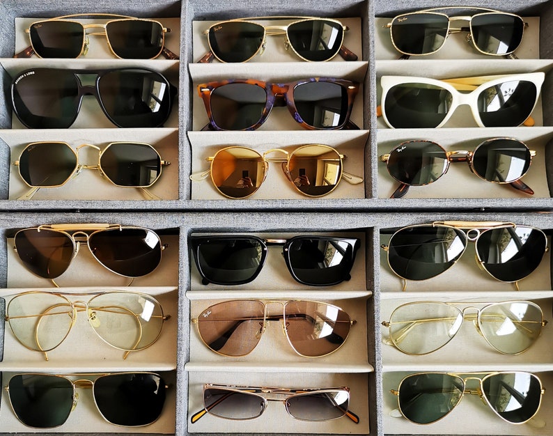 May include: A collection of sunglasses in various styles and colors, including aviator, cat-eye, and rectangular frames. The sunglasses are displayed in a gray organizer, showcasing a variety of lens tints and frame materials, such as gold and tortoiseshell.