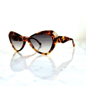 Op de afbeelding: Cat-eye zonnebril met een schildpadpatroon. De monturen zijn bruin en geel met een gouden accent op de slapen. De glazen hebben een gradiënttint, van donker naar licht. Een modieus accessoire.
