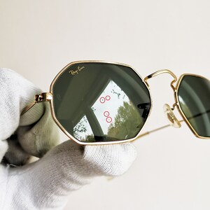 Pu&ograve; includere: Occhiali da sole con montatura dorata e lenti verdi di forma esagonale. Gli occhiali hanno il logo Ray-Ban sulla lente sinistra.