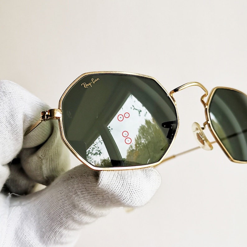 Pu&ograve; includere: Occhiali da sole con montatura dorata e lenti verdi di forma esagonale. Gli occhiali hanno il logo Ray-Ban sulla lente sinistra.