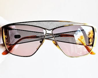 Peut inclure: Une paire de lunettes de soleil à monture dorée avec un cadre en maille noire et des verres bruns. Les lunettes ont une forme angulaire unique.