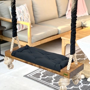 May include: A wooden swing with black rope and a black padded seat. The swing is hanging from a wooden beam and is in front of a couch and a rug.