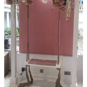 May include: A white swing with a pink cushion hangs from a white frame decorated with a floral garland of pink and white roses. The swing is in front of a pink wall.