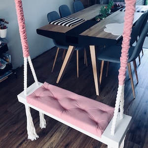 May include: A white wooden swing with a pink padded seat and white rope hanging from the ceiling. The swing is hanging in a room with a wooden floor and a table with chairs.