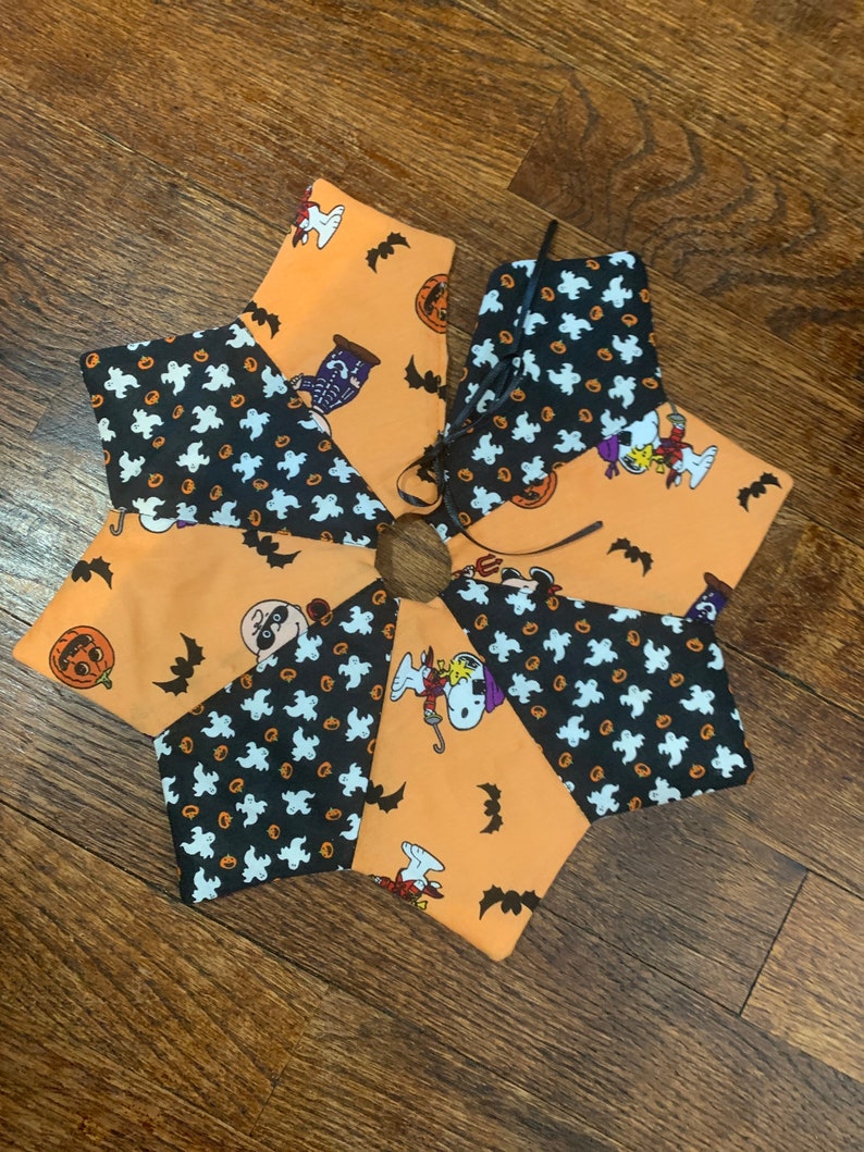 May include: A six-pointed star-shaped tree skirt with a Halloween theme. The skirt is made of orange fabric with black bats and pumpkins. The center of the skirt is black with white ghosts and pumpkins. The skirt has a black ribbon tie.