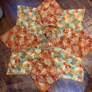 May include: A star-shaped tree skirt with a fall leaf pattern in shades of orange, brown, and green. The skirt has a brown ribbon tied in a bow in the center.
