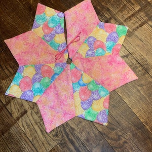 May include: A pink and yellow fabric Christmas tree skirt with a pastel seashell pattern. The skirt has a pink ribbon tied in the centre.