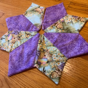 May include: A star-shaped fabric decoration with alternating panels. Some panels feature a purple floral pattern, while others display a spring scene with bunnies, daisies, and Easter eggs. The decoration is likely handmade and suitable for Easter.