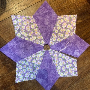 以下が含まれることがあります： 紫と白の布でできた8角形の星形。中心に穴が開いています。星形には、デイジー柄がプリントされています。