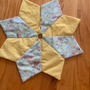 May include: A star-shaped, handmade Easter tree skirt. The skirt is made of yellow and light green fabric with a floral and bunny print. A yellow ribbon is attached to the centre. The skirt is approximately 61 cm in diameter.