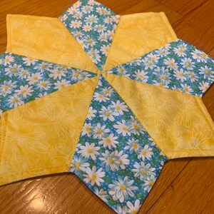 以下が含まれることがあります： 星型のキルト鍋敷き。トンボ模様の黄色い生地と白いデイジー模様の青い生地が交互に配置されています。熱いお皿を置くのに使用されます。