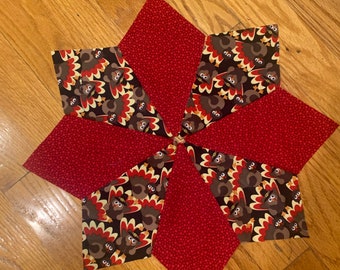 May include: A red and brown fabric patchwork quilt block with a turkey print. The block is made up of six triangles, each with a different pattern. The triangles are arranged in a star shape.