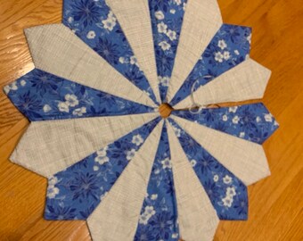 May include: A blue and white quilted Christmas tree skirt with a starburst design. The skirt is made of fabric with a floral pattern and has a white center.