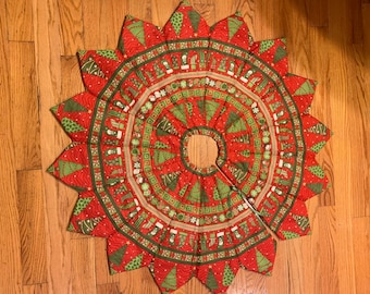 May include: A red and green Christmas tree skirt with a quilted design. The skirt features a repeating pattern of small Christmas trees, ornaments, and stockings. The skirt is made of fabric and has a scalloped edge.