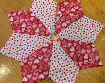 May include: A star-shaped tree skirt made of fabric with a heart pattern in shades of red, pink, and white. The skirt has a central opening and is decorated with a wooden ring and a white string.