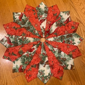 May include: A red and green Christmas tree skirt with a poinsettia and cardinal design. The skirt is made of fabric and has a starburst shape.