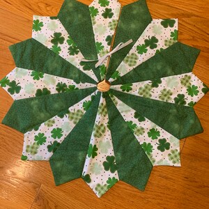 May include: A St. Patrick's Day tree skirt with a starburst design. Alternating sections of solid and patterned green fabric, featuring shamrocks and plaid patterns. A small, woven raffia-like circle is at the center, with a white ribbon tied to it.