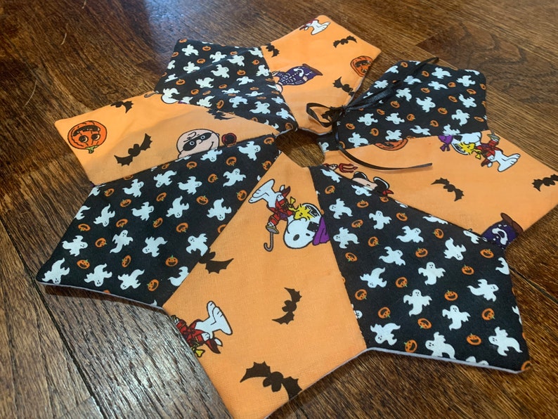 May include: A multi-colored fabric tree skirt with a black and white ghost and pumpkin pattern, an orange and black Peanuts pattern, and a black and white Peanuts pattern. The skirt has a black ribbon tied around the center.