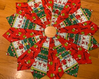May include: A red and green Christmas tree skirt with a Grinch theme. The skirt is made of fabric with a repeating pattern of the Grinch's face and the words "Merry Grinchmas".