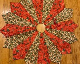 May include: A star-shaped Christmas tree skirt with a wooden center. The skirt is made of fabric with a pattern of red poinsettias and green holly leaves on a cream background. A red ribbon is tied on one of the points.