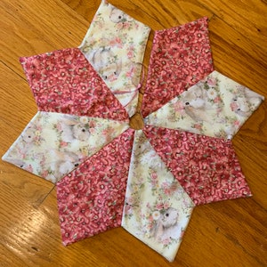 May include: A star-shaped fabric item with alternating segments of floral and bunny print fabric. The floral fabric is a deep red with small flowers, while the bunny print has a cream background with bunnies and roses. A pink ribbon is attached at the centre.