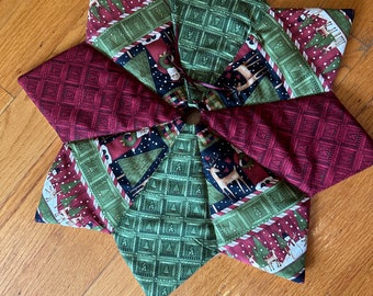 May include: A star-shaped Christmas tree skirt made of fabric. The skirt features alternating sections of patterned fabric in shades of green, burgundy, and navy. The patterns include Christmas trees, reindeer, and geometric designs. A brown button is in the center.