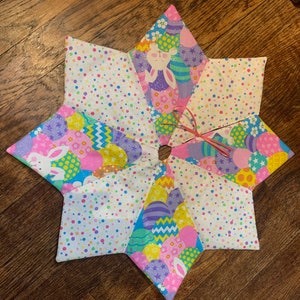 May include: A star-shaped Easter tree skirt with a central opening. The skirt is made of fabric panels featuring Easter eggs, bunnies, and colorful polka dots. A pink ribbon is tied near the centre.
