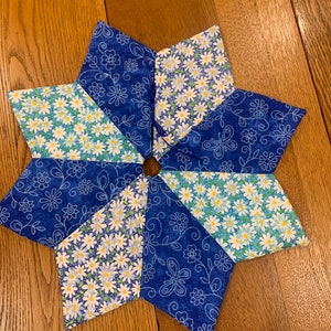 以下が含まれることがあります： 青い花柄とデイジー柄の生地を交互に使用した、星型のキルト鍋敷き。熱から表面を保護するように設計されており、キッチンやダイニングエリアに装飾的なタッチを加えます。直径は約25cmです。