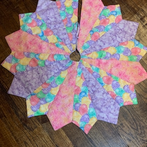 May include: A colorful patchwork tree skirt with a pastel pink, yellow, and purple design featuring a pattern of eggs. The skirt has a circular shape with a hole in the center for a tree trunk.