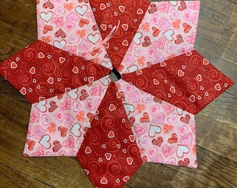 May include: A red and pink fabric tree skirt with a heart pattern. The skirt is made of multiple pieces of fabric sewn together to form a star shape.