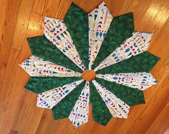 May include: A handmade Christmas tree skirt with a starburst design. Alternating green and patterned fabric panels radiate from a central wooden circle. The patterned fabric features festive icons like trees, stars, and mittens. A green ribbon is attached.