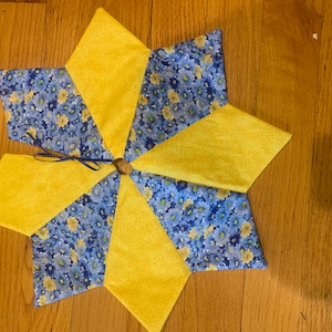 以下が含まれることがあります： 星型のツリースカートで、黄色と青の花柄の生地が交互に配置されています。中央には木製のリングと青いリボンが付いています。スカートはクリスマスツリーの根元に置くようにデザインされています。